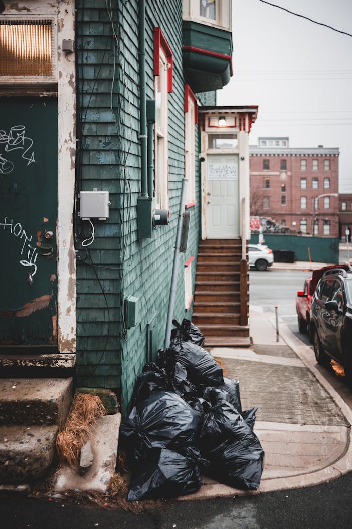 Black plastic garbage bags placed on sidewalk near colorful residential house with shabby stairs on street with buildings and cars
